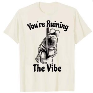 Puede incluir: Camiseta color crema con una ilustraci&oacute;n en blanco y negro de una rana con gafas de sol y un palo. El texto "You're Ruining The Vibe" est&aacute; impreso encima y debajo del gr&aacute;fico de la rana en una fuente vintage.