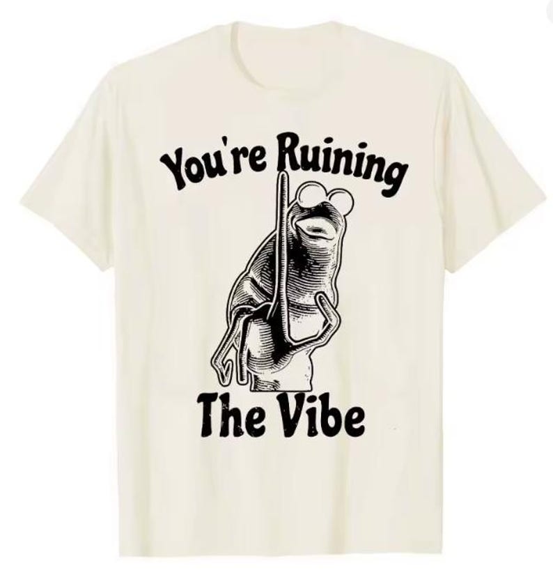 Puede incluir: Camiseta color crema con una ilustraci&oacute;n en blanco y negro de una rana con gafas de sol y un palo. El texto "You're Ruining The Vibe" est&aacute; impreso encima y debajo del gr&aacute;fico de la rana en una fuente vintage.