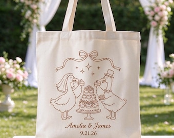 May include: A cream-colored canvas tote bag featuring a brown line-art illustration of two geese, one wearing a veil and the other a top hat, standing by a tiered wedding cake. The text reads Amelia & James and the date 9.21.26.