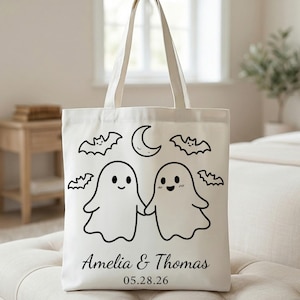 May include: A cream-colored canvas tote bag featuring a black line art graphic of two ghosts holding hands, surrounded by four small bats and a crescent moon. Below the ghosts, the text reads Amelia & Thomas followed by the date 05.28.26.