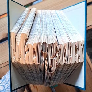 Può includere: Un libro con pagine piegate che formano il nome Margaux. Il libro ha una copertina blu scuro e le pagine sono color crema con testo visibile. Le pagine sono piegate e curvate con precisione per creare le lettere tridimensionali del nome.