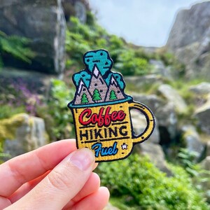 May include: A yellow patch in the shape of a coffee mug with the words "Coffee Hiking Fuel". The mug design includes a mountain scene with green trees and white peaks. The patch is held against a backdrop of rocks and greenery.