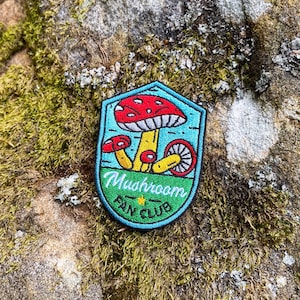 May include: An embroidered patch featuring a red and white mushroom, with the text "Mushroom Fan Club" in blue and green.