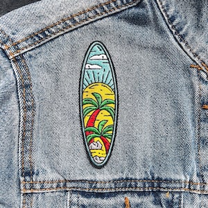 Könnte beinhalten: Eine blaue Jeansjacke mit einem bunten gestickten Aufnäher einer Surfboard mit einer Palme und einer Sonne.