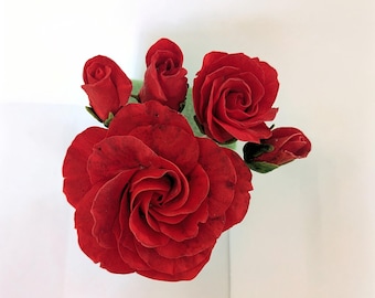 Op de afbeelding: Een tros van vijf dieprode rozen, waaronder één grote volledig bloeiende bloem, twee kleinere half geopende bloesems en twee dichte rozenknoppen, allemaal met gelaagde bloemblaadjes met subtiele donkere schaduwen en groene kelkblaadjes.