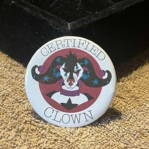 May include: A circular badge with the text "CERTIFIED CLOWN". The design features a cartoon clown with black hair, a red nose, and a purple collar. The background is white with a red and purple border. The badge is on a textured surface.