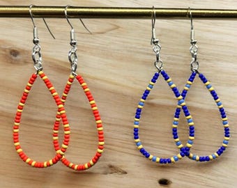 May include: Two pairs of beaded teardrop earrings. One pair is red and yellow, the other is blue and yellow. The earrings are suspended from silver-colored hooks. The background is a light brown wooden surface.