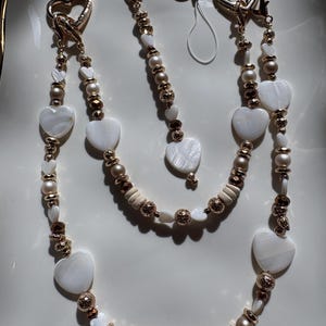 May include: A set of three beaded accessories with heart-shaped and round white beads, interspersed with gold and bronze-colored beads. The accessories include a phone charm and two lanyards with heart-shaped clasps.
