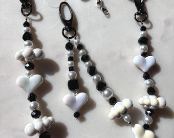 May include: A beaded phone charm with black, white, and iridescent beads. The charm features white heart and cloud-shaped beads, along with black square and round beads. The charm has black metal clasps and rings.