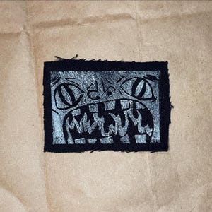 Peut inclure: Un écusson rectangulaire noir avec une illustration argentée d'un visage hurlant. Le dessin présente de grands yeux et une bouche large et dentée, avec un style vieilli et dessiné à la main. L'écusson est probablement destiné aux vêtements ou aux accessoires.