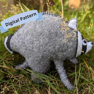 May include: A gray and white felt raccoon plush toy featuring a black eye mask and a striped tail. A light blue banner with the text Digital Pattern is attached to the top left. The raccoon has visible stitched seams along its edges.