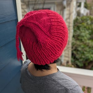 May include: A vibrant red knit hat with a tassel detail. The hat is made of a thick, textured yarn and has a rounded shape. The hat is worn by a person wearing a gray t-shirt.