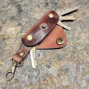 Peut inclure: Un porte-clés en cuir marron avec une fermeture à pression en métal et un anneau. Le porte-clés a un rabat qui recouvre les clés et un petit bouton en métal sur le rabat. Les clés sont visibles à l'intérieur du porte-clés.