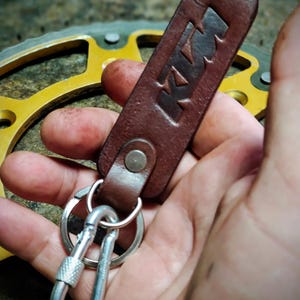 Può includere: Un portachiavi in ​​cuoio marrone con un moschettone argentato e la parola "KTM" impressa sul cuoio.