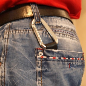 Könnte beinhalten: Nahaufnahme einer Gesäßtasche mit einem silbernen Karabiner, der an einem schwarzen Gürtel befestigt ist. Die Jeans sind blau mit einem roten, weißen und blauen Streifenmuster.