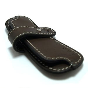 May include: A brown leather case with white stitching and a silver button. The case is designed to hold a small knife or other tool.