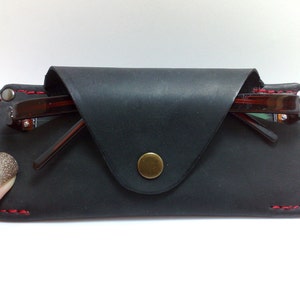 May include: Black leather glasses case with red stitching and a brass snap closure. The case holds a pair of brown-framed eyeglasses. The case is rectangular with a flap closure.
