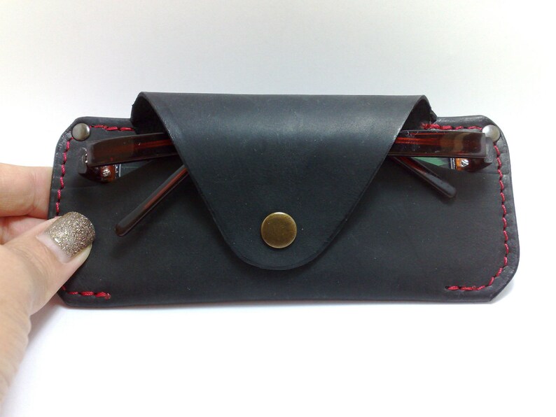 May include: Black leather glasses case with red stitching and a brass snap closure. The case holds a pair of brown-framed eyeglasses. The case is rectangular with a flap closure.