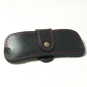 May include: A black leather case with red stitching and a brass button closure. The case is designed to hold a small item, such as a knife or a key.
