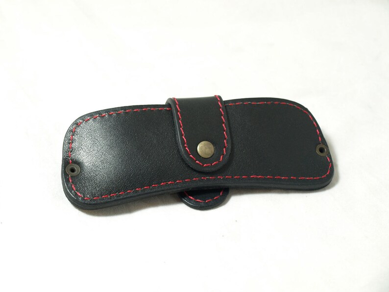 May include: A black leather case with red stitching and a brass button closure. The case is designed to hold a small item, such as a knife or a key.