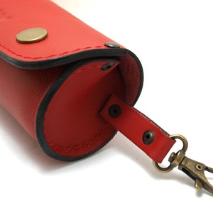 May include: Red leather eyeglasses case with a black stitching detail and a brass clasp. The case has a strap with a brass snap hook.