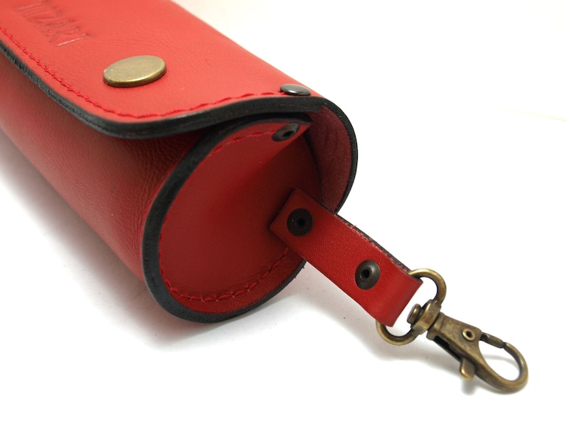 May include: Red leather eyeglasses case with a black stitching detail and a brass clasp. The case has a strap with a brass snap hook.