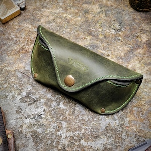 Puede incluir: Un estuche de gafas de sol de cuero verde con la palabra "VAIDO" grabada en relieve en la parte delantera. El estuche tiene un cierre de bot&oacute;n y costuras verdes. El estuche est&aacute; sobre una superficie texturizada.