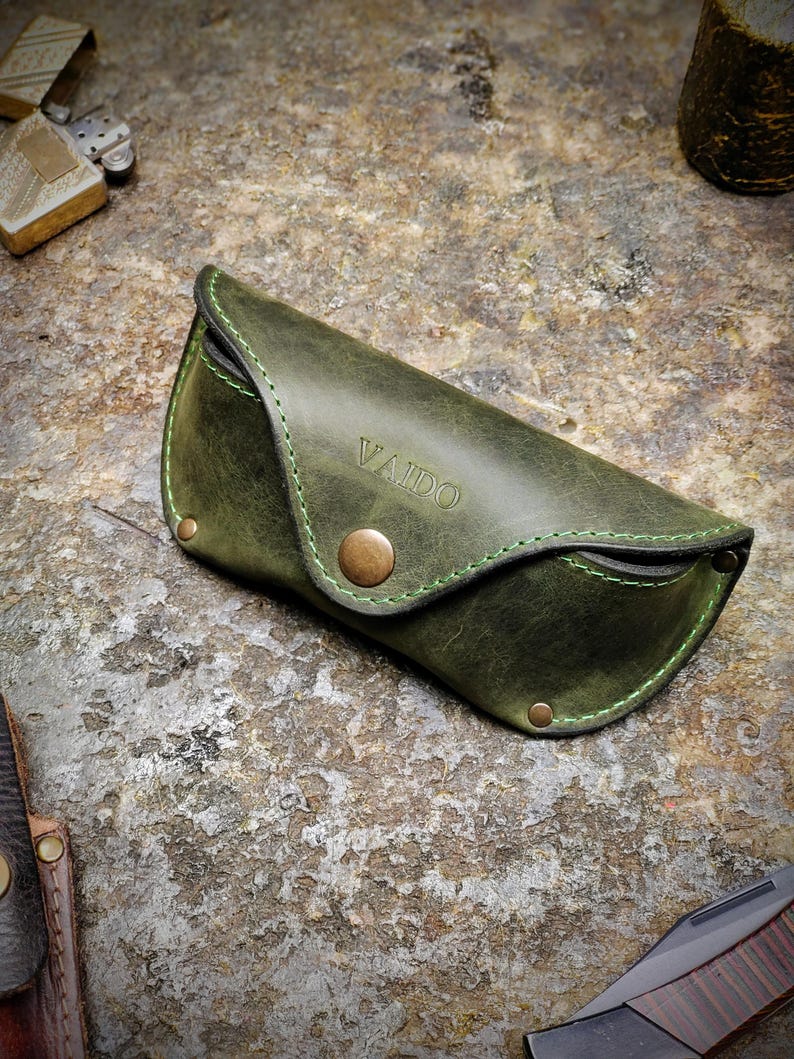Puede incluir: Un estuche de gafas de sol de cuero verde con la palabra "VAIDO" grabada en relieve en la parte delantera. El estuche tiene un cierre de bot&oacute;n y costuras verdes. El estuche est&aacute; sobre una superficie texturizada.