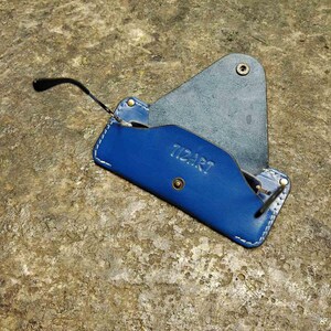 May include: A blue leather glasses case with white stitching and the word "TIZART" embossed on the front. The case is open, revealing a pair of black-framed glasses. The case is on a textured, brown surface.