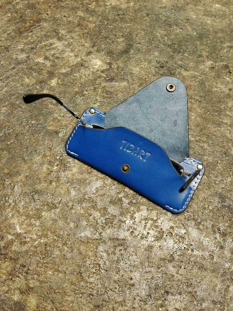 May include: A blue leather glasses case with white stitching and the word "TIZART" embossed on the front. The case is open, revealing a pair of black-framed glasses. The case is on a textured, brown surface.