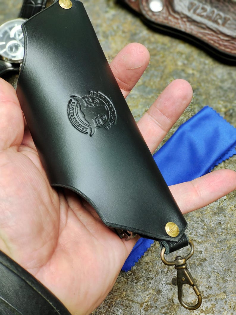 Puede incluir: Llavero de cuero negro con cierre y remaches de lat&oacute;n. El llavero presenta un emblema en relieve con el texto "LEATHER CRAFTS" y un dise&ntilde;o de cabeza de toro. Se ve un pa&ntilde;o de microfibra azul.