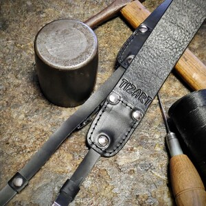 May include: Close-up of a black leather camera strap with the embossed text "TIZART". The strap features silver rivets and a buckle. A metal hammer, a mallet, and a leather needle are also visible.