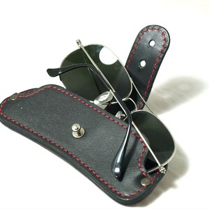 May include: A black leather sunglasses case with red stitching and a silver snap closure. The case is open and a pair of sunglasses are inside.