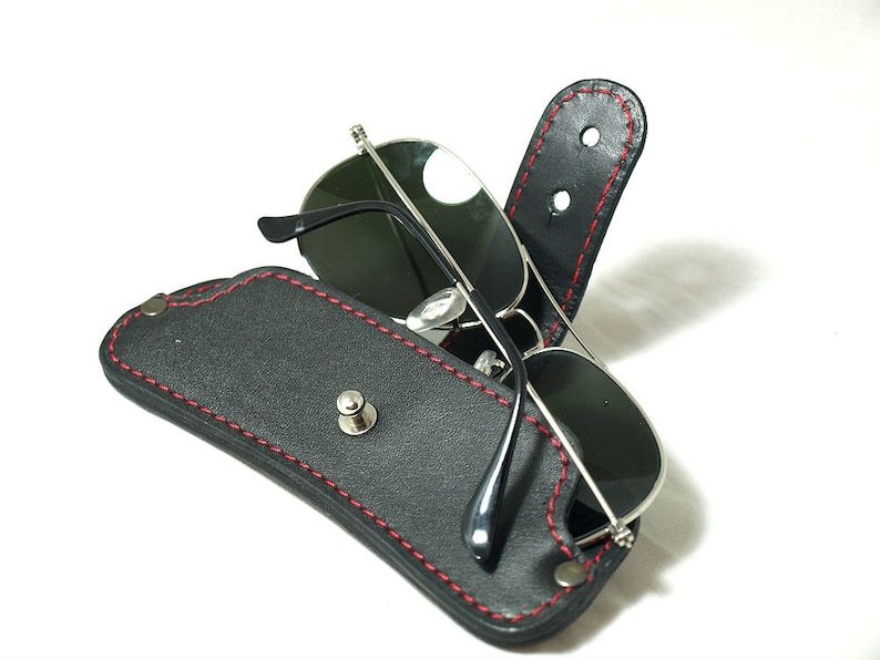 May include: A black leather sunglasses case with red stitching and a silver snap closure. The case is open and a pair of sunglasses are inside.