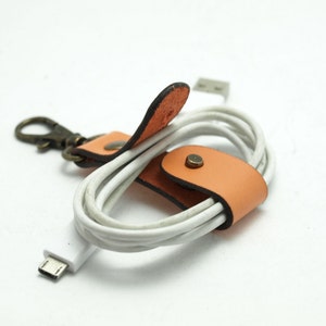 May include: A white charging cable with a leather cord organiser. The organiser is tan with a brown trim and a metal clasp.