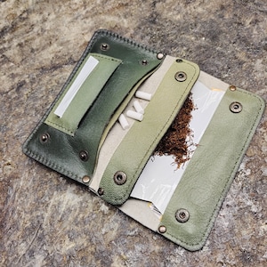 Puede incluir: Una bolsa de tabaco de cuero verde con cierre de solapa y un compartimento separado para filtros. La bolsa está abierta, revelando tabaco y filtros en su interior.