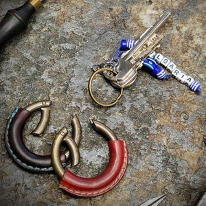 Pode incluir: Close-up de chaveiros artesanais de couro e metal em tons de vermelho e castanho. Um conjunto de chaves com um chaveiro de contas "BULGARIA" também é visível. Os chaveiros estão sobre uma superfície texturizada semelhante a pedra.