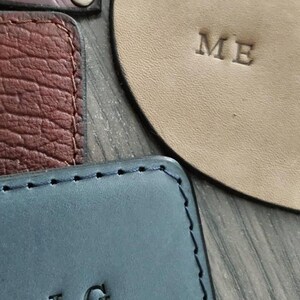 Puede incluir: Primer plano de varios art&iacute;culos de cuero, incluyendo una pieza rectangular azul con "LG" en relieve, una pieza beige con "ME", y otras piezas de cuero texturizado en marr&oacute;n y burdeos. Los art&iacute;culos parecen hechos a mano.