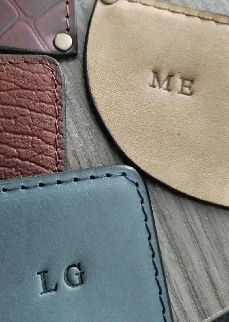 Puede incluir: Primer plano de varios art&iacute;culos de cuero, incluyendo una pieza rectangular azul con "LG" en relieve, una pieza beige con "ME", y otras piezas de cuero texturizado en marr&oacute;n y burdeos. Los art&iacute;culos parecen hechos a mano.