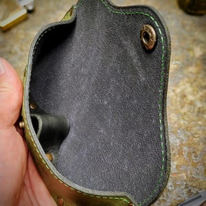 Puede incluir: Una funda de cuero hecha a mano con un exterior verde y marr&oacute;n y un interior gris oscuro. La funda presenta costuras verdes y remaches de lat&oacute;n. El dise&ntilde;o es para llevar un objeto peque&ntilde;o.