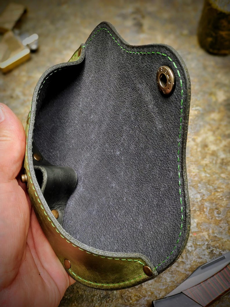Puede incluir: Una funda de cuero hecha a mano con un exterior verde y marr&oacute;n y un interior gris oscuro. La funda presenta costuras verdes y remaches de lat&oacute;n. El dise&ntilde;o es para llevar un objeto peque&ntilde;o.