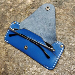 May include: A blue leather glasses case with white stitching and a snap closure. The case is open, revealing a pair of black-framed glasses. The case is on a textured, brown surface.