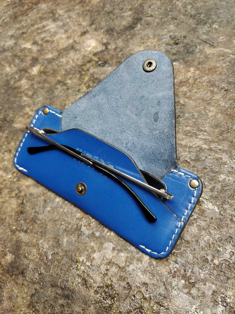 May include: A blue leather glasses case with white stitching and a snap closure. The case is open, revealing a pair of black-framed glasses. The case is on a textured, brown surface.