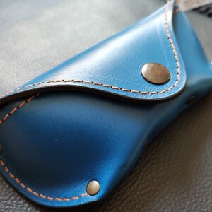 Puede incluir: Un estuche para gafas de cuero azul con costuras naranjas y un cierre de bot&oacute;n a presi&oacute;n de color bronce. El estuche es rectangular con la parte superior e inferior curvadas.