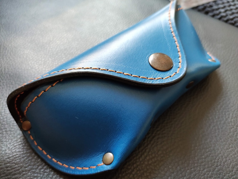 Puede incluir: Un estuche para gafas de cuero azul con costuras naranjas y un cierre de bot&oacute;n a presi&oacute;n de color bronce. El estuche es rectangular con la parte superior e inferior curvadas.
