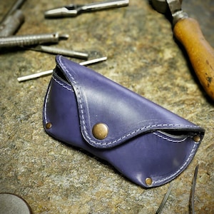 Puede incluir: Un estuche de gafas de cuero morado hecho a mano, con costuras blancas y cierre de bot&oacute;n de lat&oacute;n. El estuche est&aacute; rodeado de varias herramientas met&aacute;licas, lo que sugiere un taller. Dise&ntilde;ado para proteger gafas.