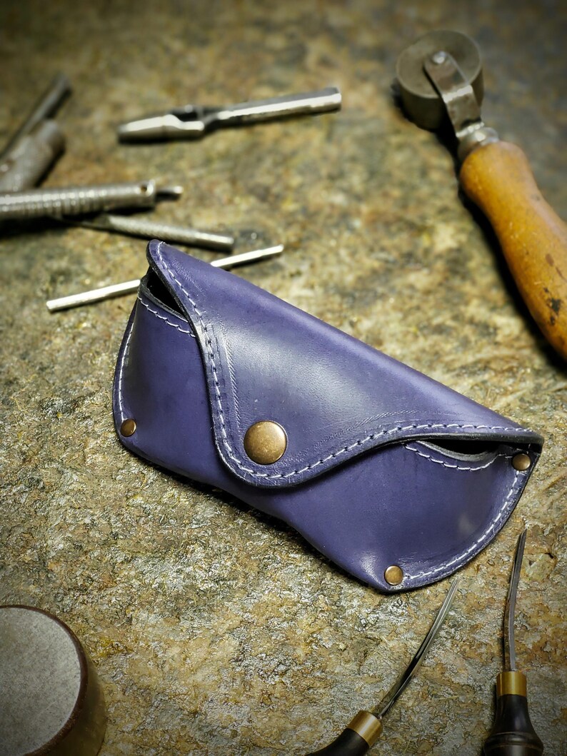 Puede incluir: Un estuche de gafas de cuero morado hecho a mano, con costuras blancas y cierre de bot&oacute;n de lat&oacute;n. El estuche est&aacute; rodeado de varias herramientas met&aacute;licas, lo que sugiere un taller. Dise&ntilde;ado para proteger gafas.