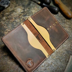 Pode incluir: Uma carteira de couro castanho artesanal com interior e detalhes em bege claro. A carteira apresenta uma borda costurada, rebites de latão e um logotipo estampado. O design inclui um slot para cartão e um bolso para dinheiro.