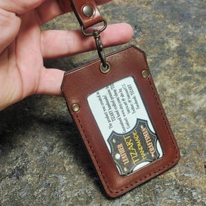 May include: A brown leather ID holder with a matching strap and metal clasp. The holder features a clear window for displaying an ID card and the text "TIZART LEATHER HANDMADE CRAFTSMAN".