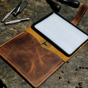 May include: A brown leather phone case with a black frame, open to reveal a white interior. The case has a textured finish and a stitched border. Small tools and hardware are scattered around the case.